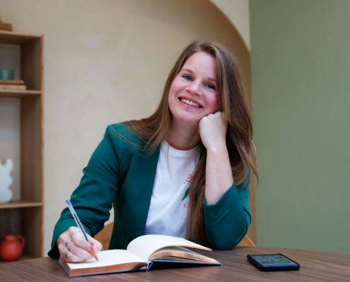 Mirjam de Nijs, familiepsycholoog, zittend aan tafel met een notitieboekje. Ze heeft haar hand onder haar kin en kijkt de camera in.